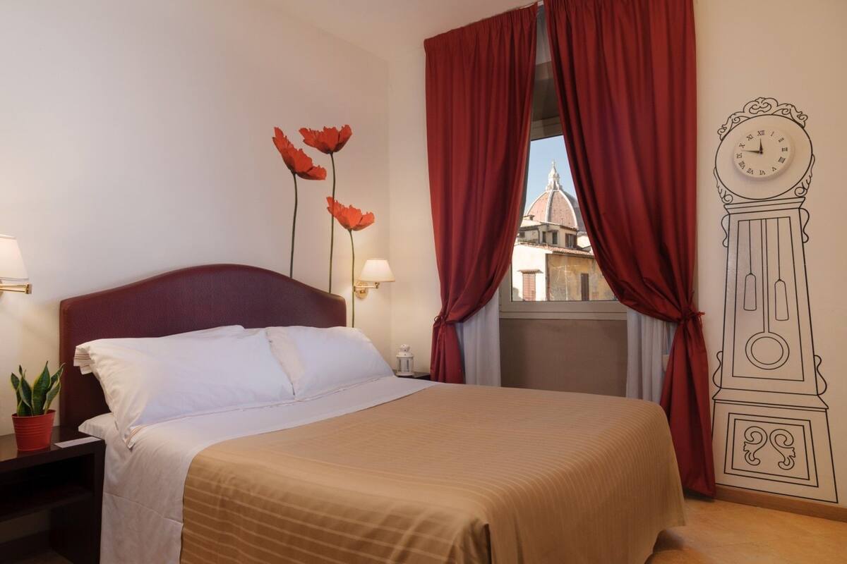 Ein Hotelzimmer mit Bett, rotem Vorhang, Wanduhr und Blick auf ein Gebäude durch das Fenster.