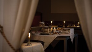 Uma sala de jantar com pouca luz, com velas, uma mesa posta com uma caixa de chá, potes e um prato de doces.