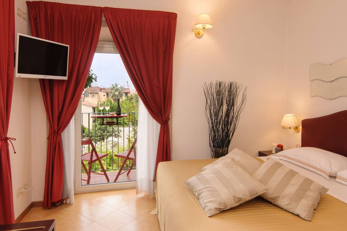 Ein Hotelzimmer mit Bett, roten Vorhängen, einem Fernseher, einem Balkon mit Stuhl und Blick auf Gebäude.