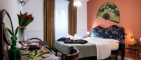 A hotel room with a bed, a desk with a vase of flowers, a chair, a table, a fan, and a wall-mounted artwork.