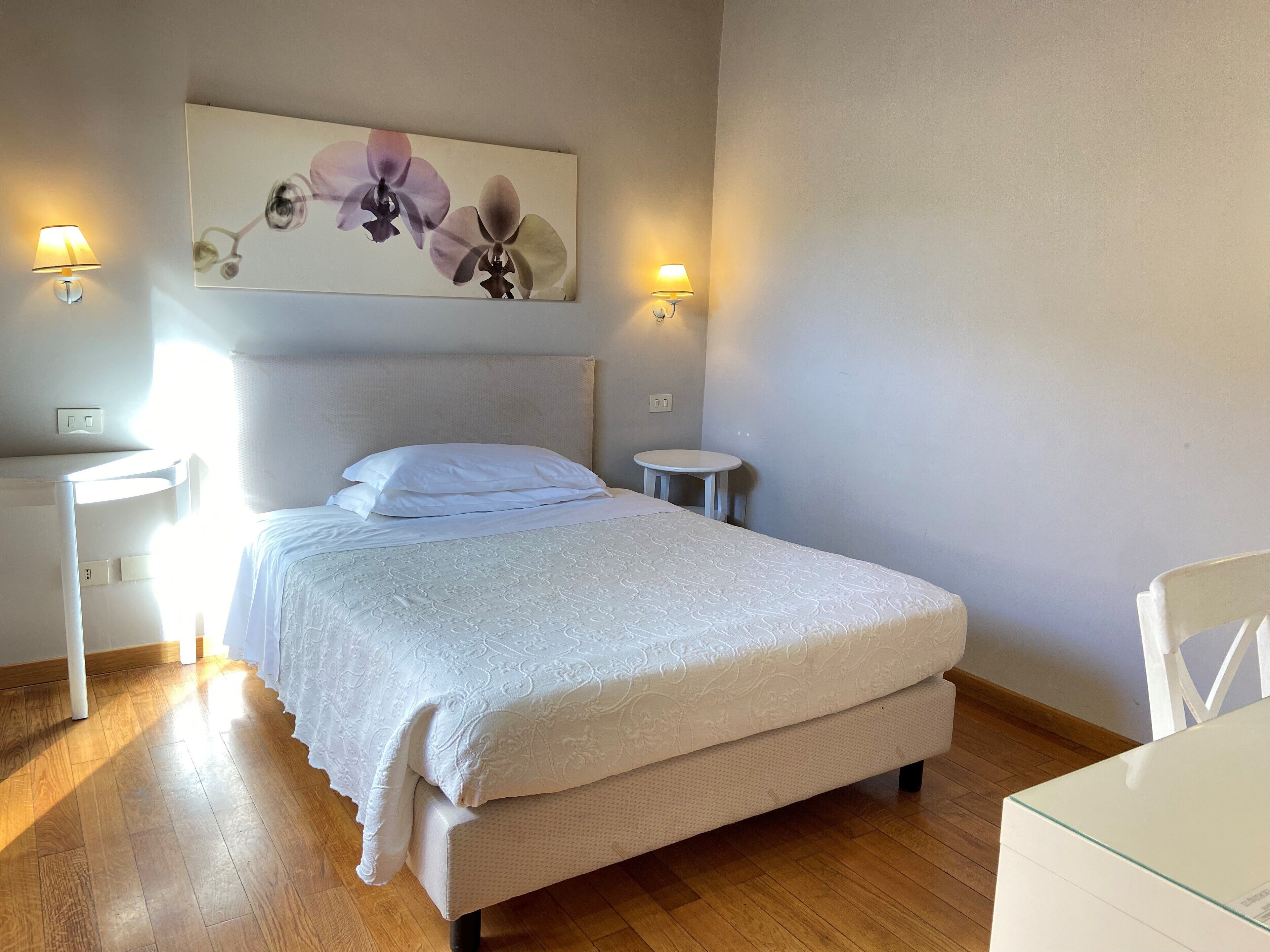 Cama bem arrumada com edredom branco, uma pequena mesa de cabeceira, luminária presa à parede e um quadro de orquídeas sobre a cama.