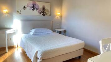 Cama bem arrumada com edredom branco, uma pequena mesa de cabeceira, luminária presa à parede e um quadro de orquídeas sobre a cama.