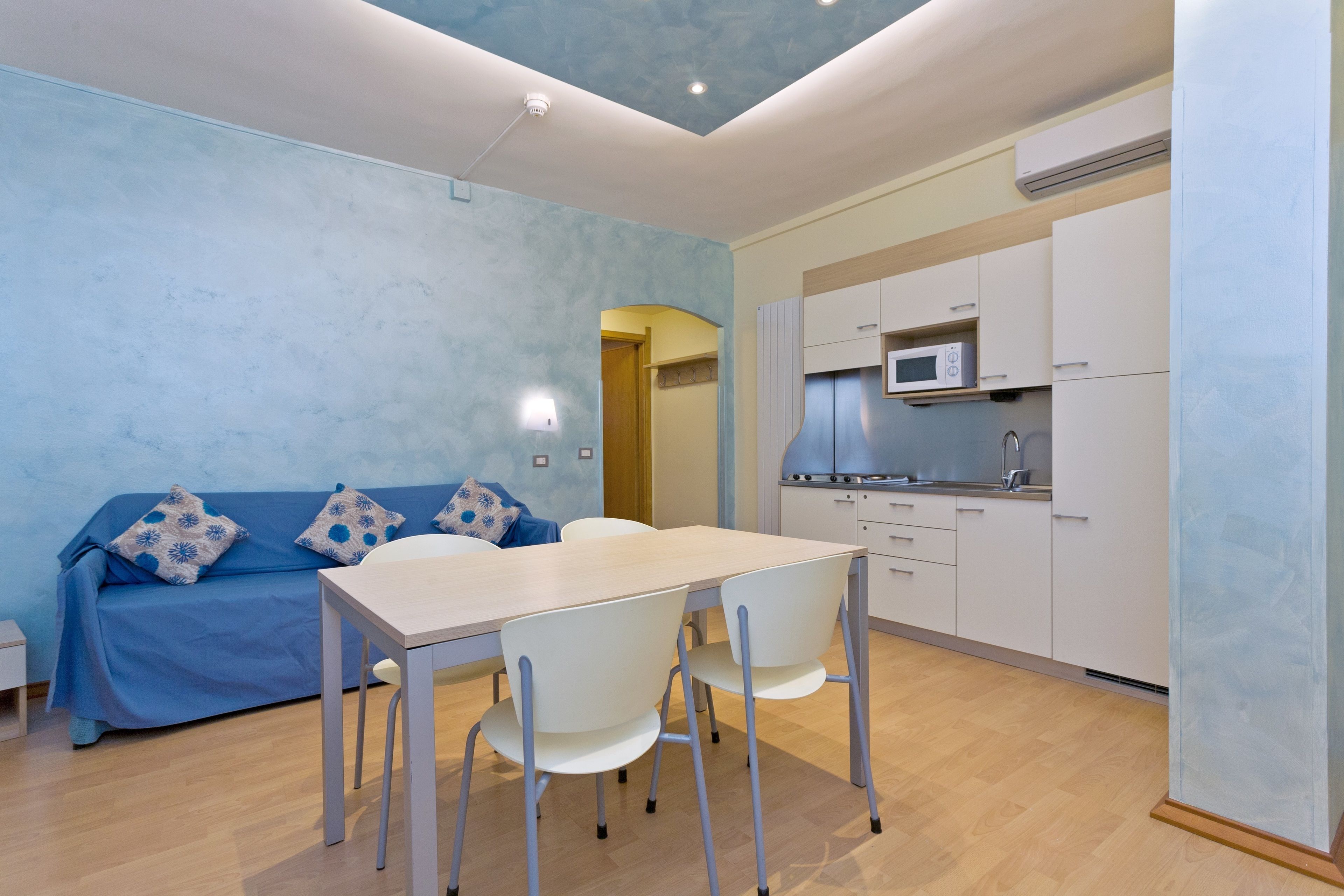Une cuisine moderne avec des meubles blancs et un four à micro-ondes, un coin repas avec une table et des chaises, et un canapé bleu agrémenté de coussins à motifs.