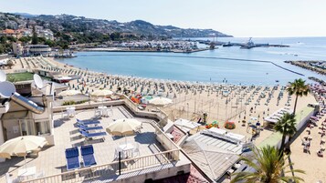 Un complejo turístico en la playa con tumbonas, sombrillas y vistas al mar y al puerto deportivo.