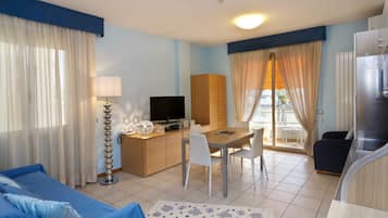 Una habitación de hotel con sofá, una mesa de comedor con sillas, televisor, armario y kitchenette.