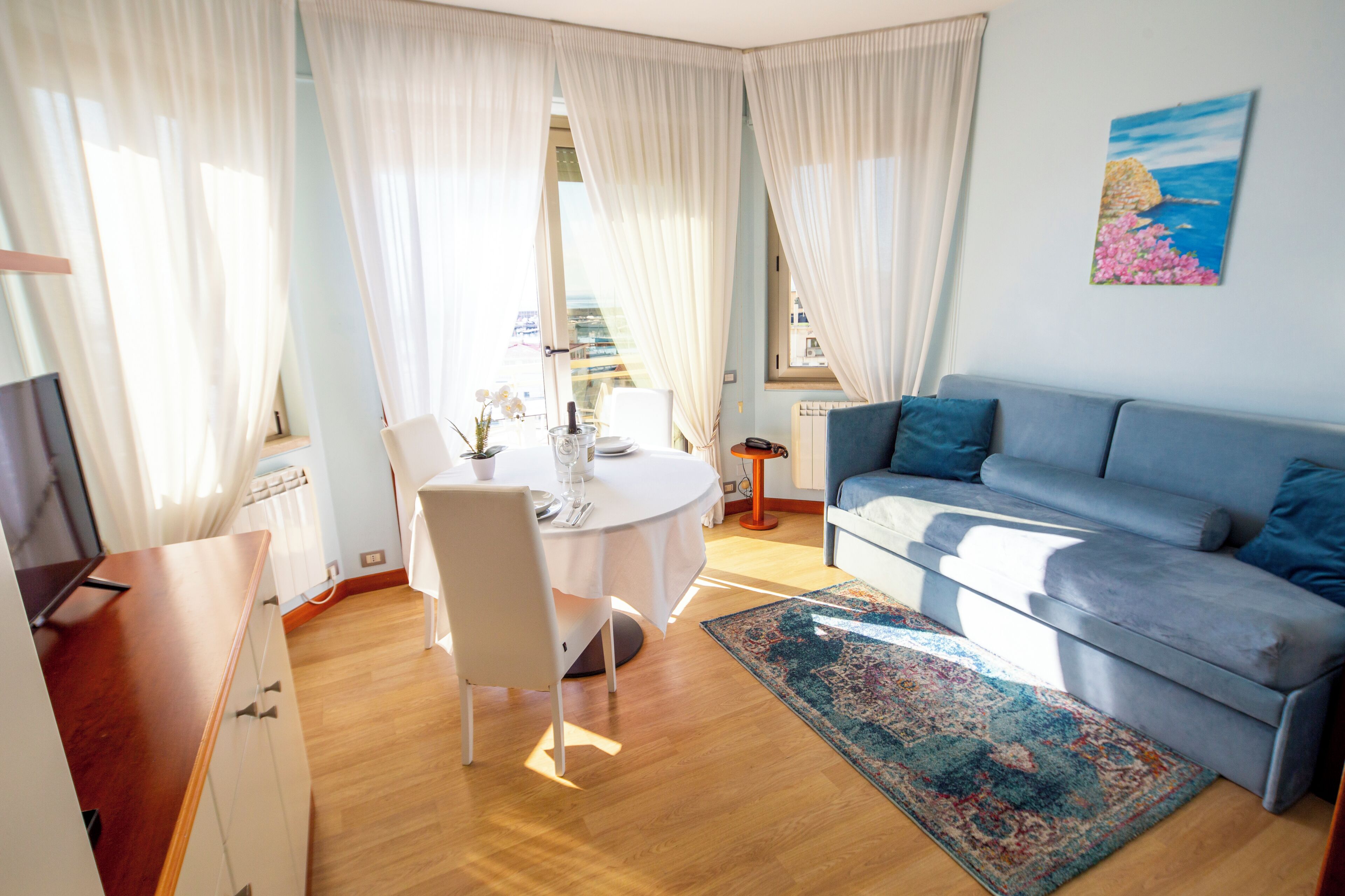 A living room with a blue sofa, a round table with chairs, and a large window with curtains.
