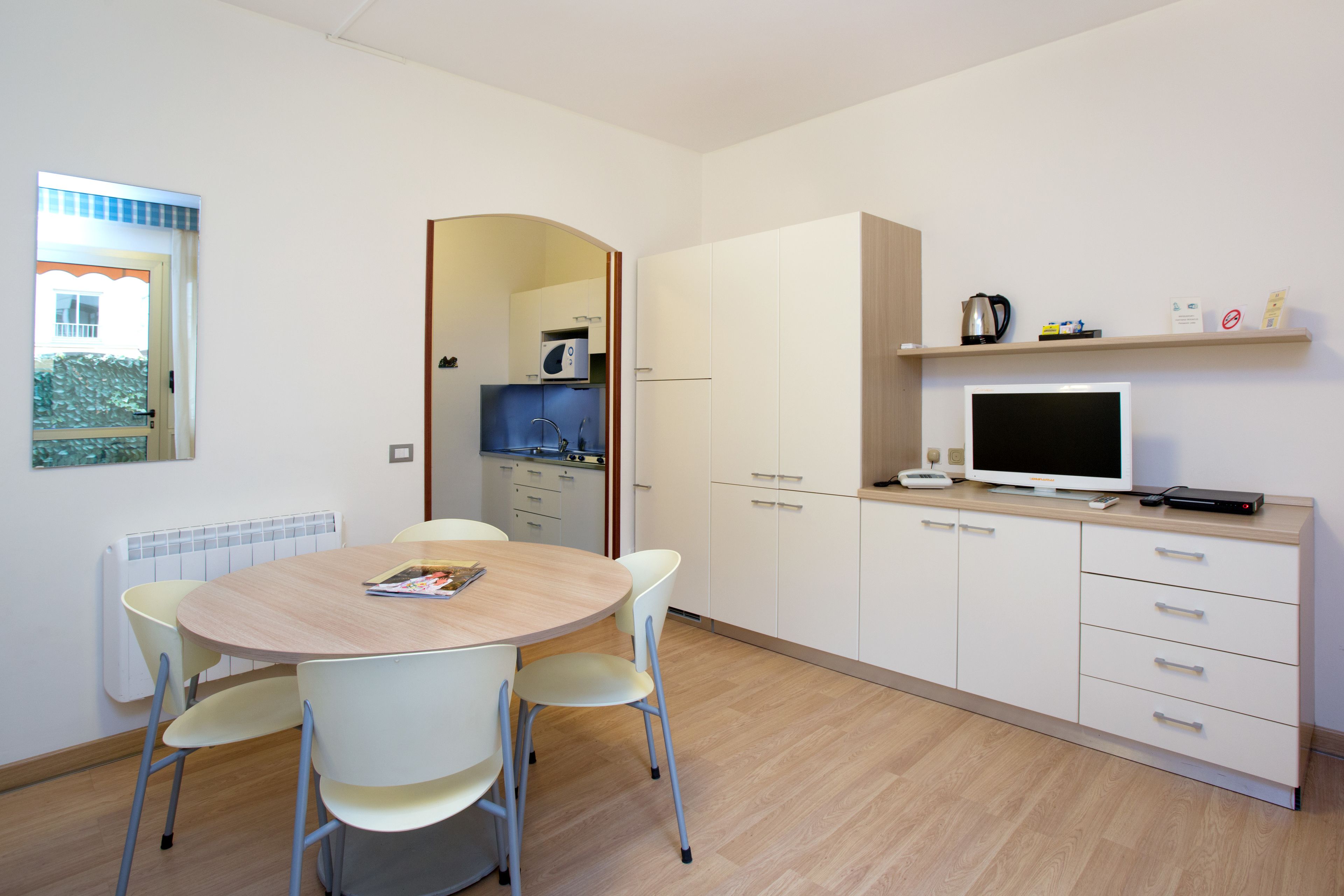 Una cocina moderna con armarios blancos, microondas y cafetera. Hay una mesa de comedor con sillas y una ventana con vistas a un edificio.