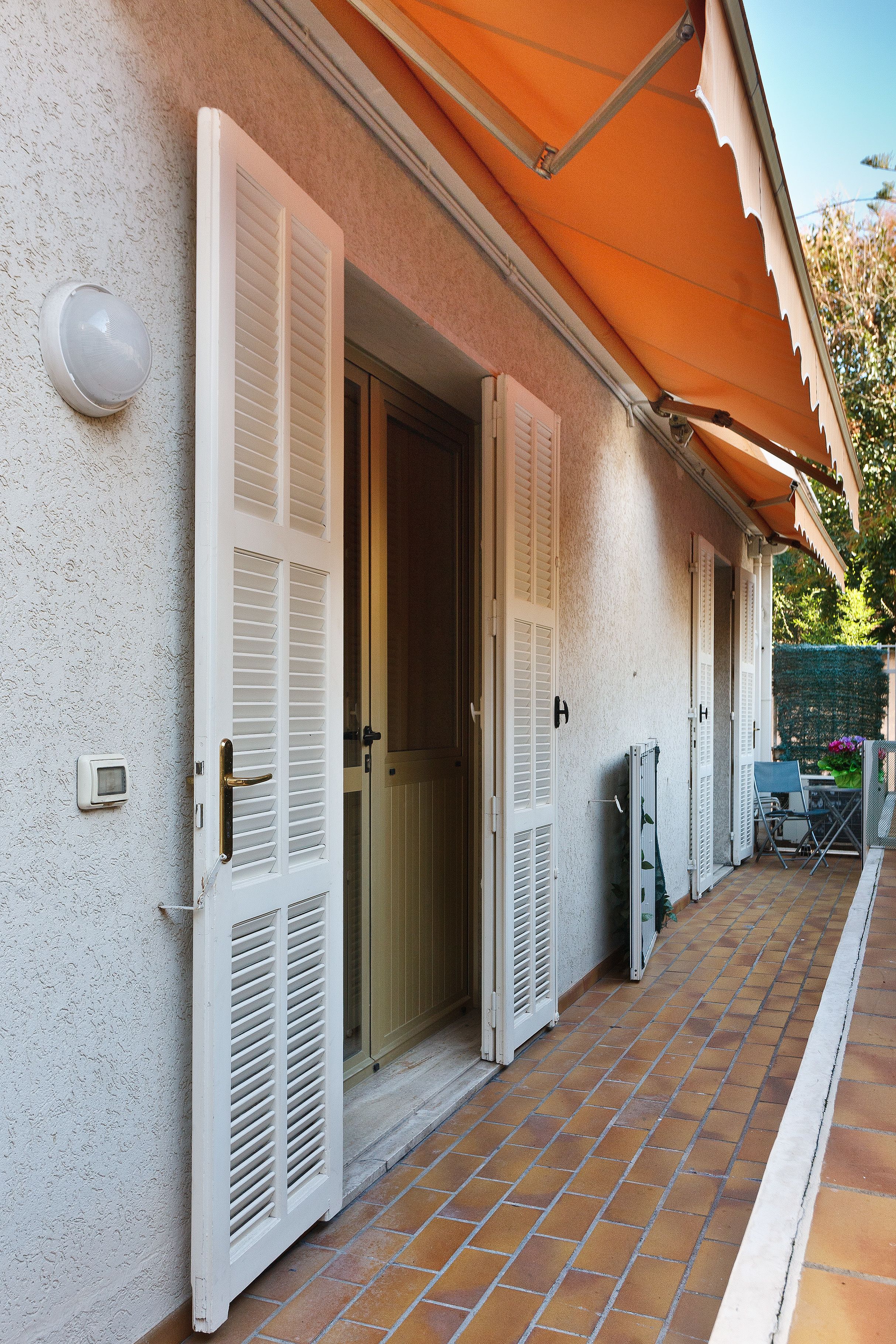Ein Haus mit weißen Fensterläden, einer gefliesten Terrasse und einer kleinen Sitzgelegenheit im Freien.