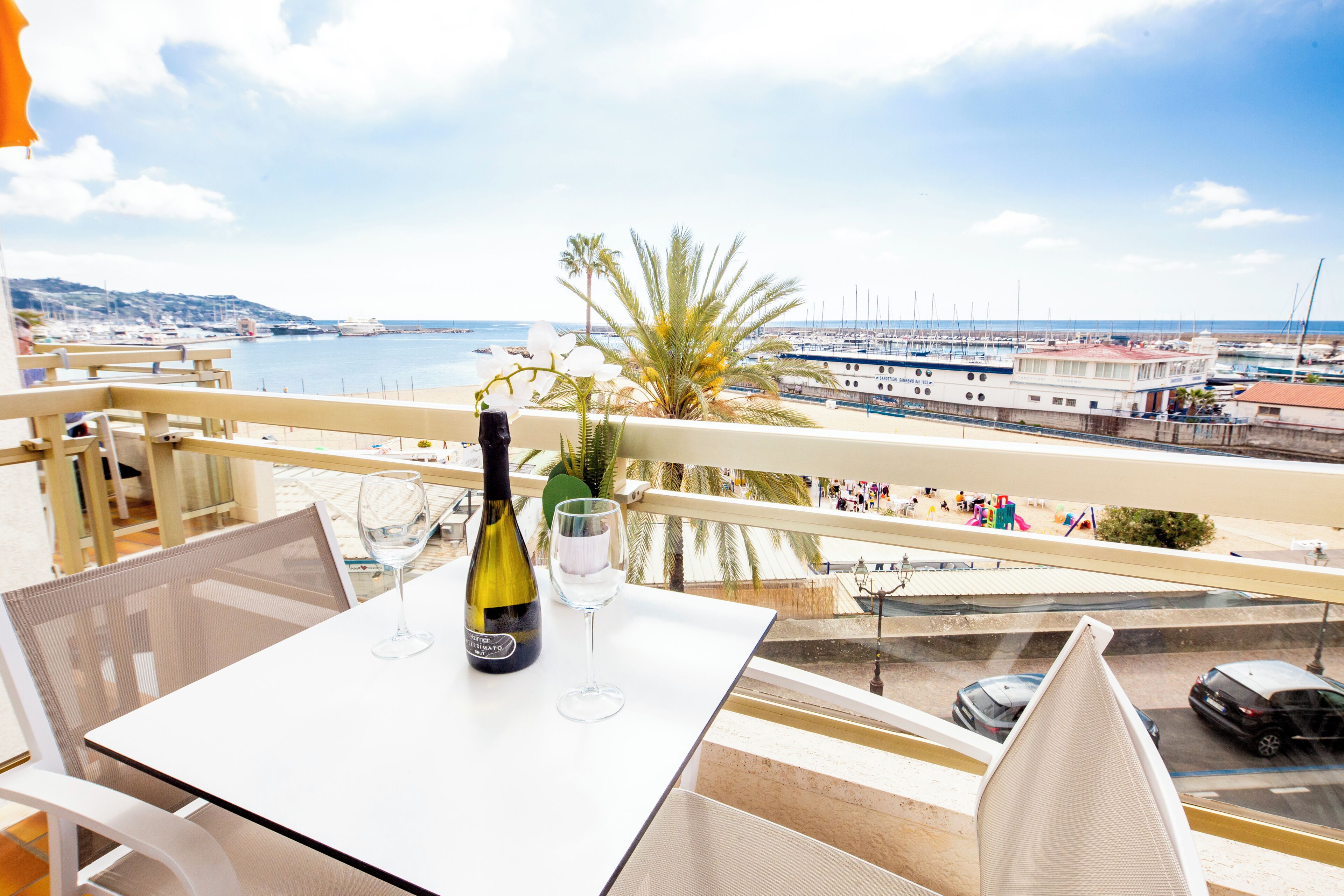 Un balcón con una mesa, una botella de vino y una copa, con vistas a un puerto deportivo con barcos y una costa lejana.