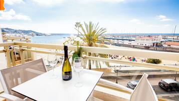 Un balcón con una mesa, una botella de vino y una copa, con vistas a un puerto deportivo con barcos y una costa lejana.