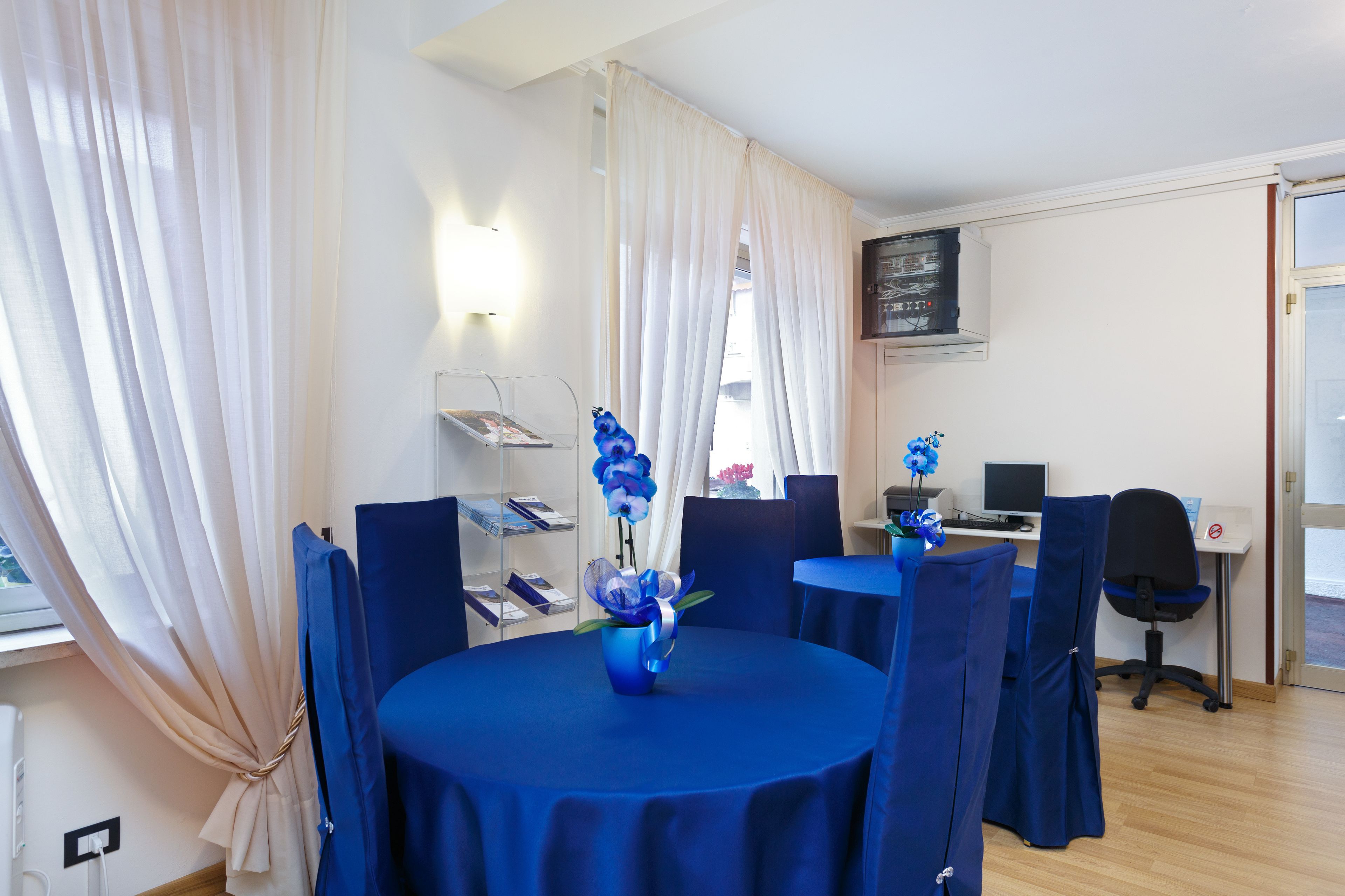 A room with a round table covered in blue tablecloth, a vase with flowers, and blue chairs. There is a desk with a computer and a chair.