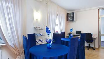 A room with a round table covered in blue tablecloth, a vase with flowers, and blue chairs. There is a desk with a computer and a chair.