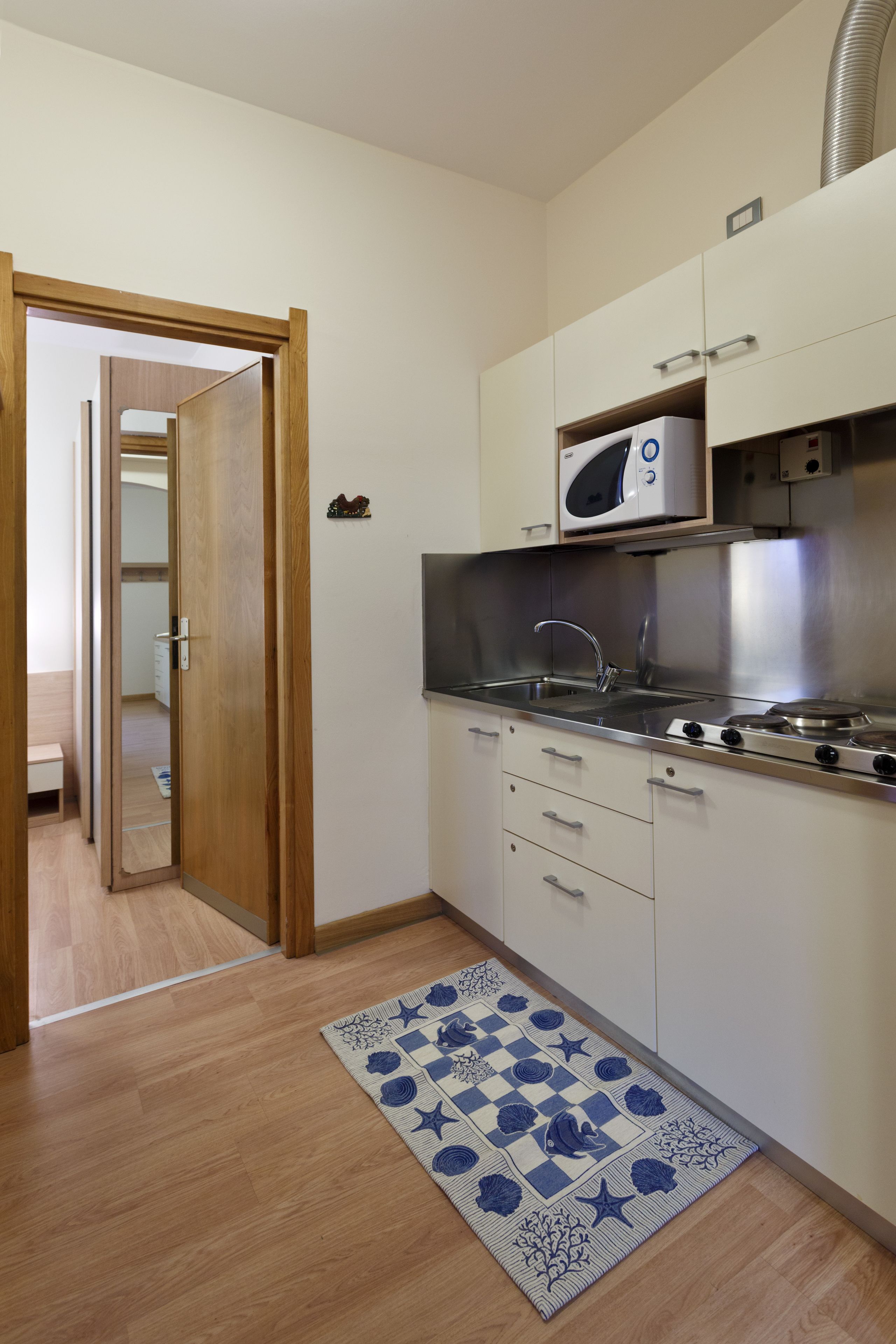 A compact kitchen with a microwave, stove, and sink. There is a wooden door leading to another room. A blue and white patterned rug is on the floor.