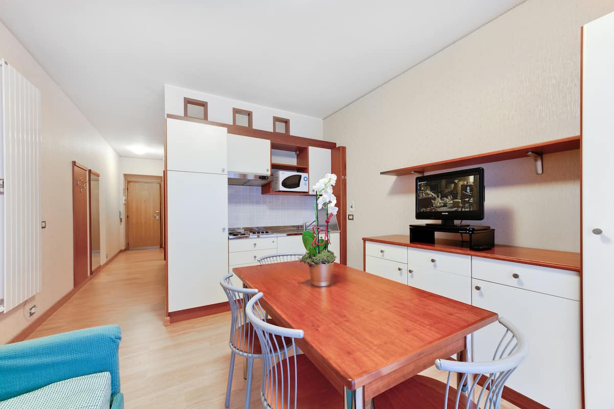 A compact kitchen with white cabinets, a wooden dining table, and a television on a wooden shelf.