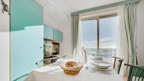 Un comedor con una mesa dispuesta para cuatro personas, un cesto de pan y vista al mar a través de un ventanal grande.