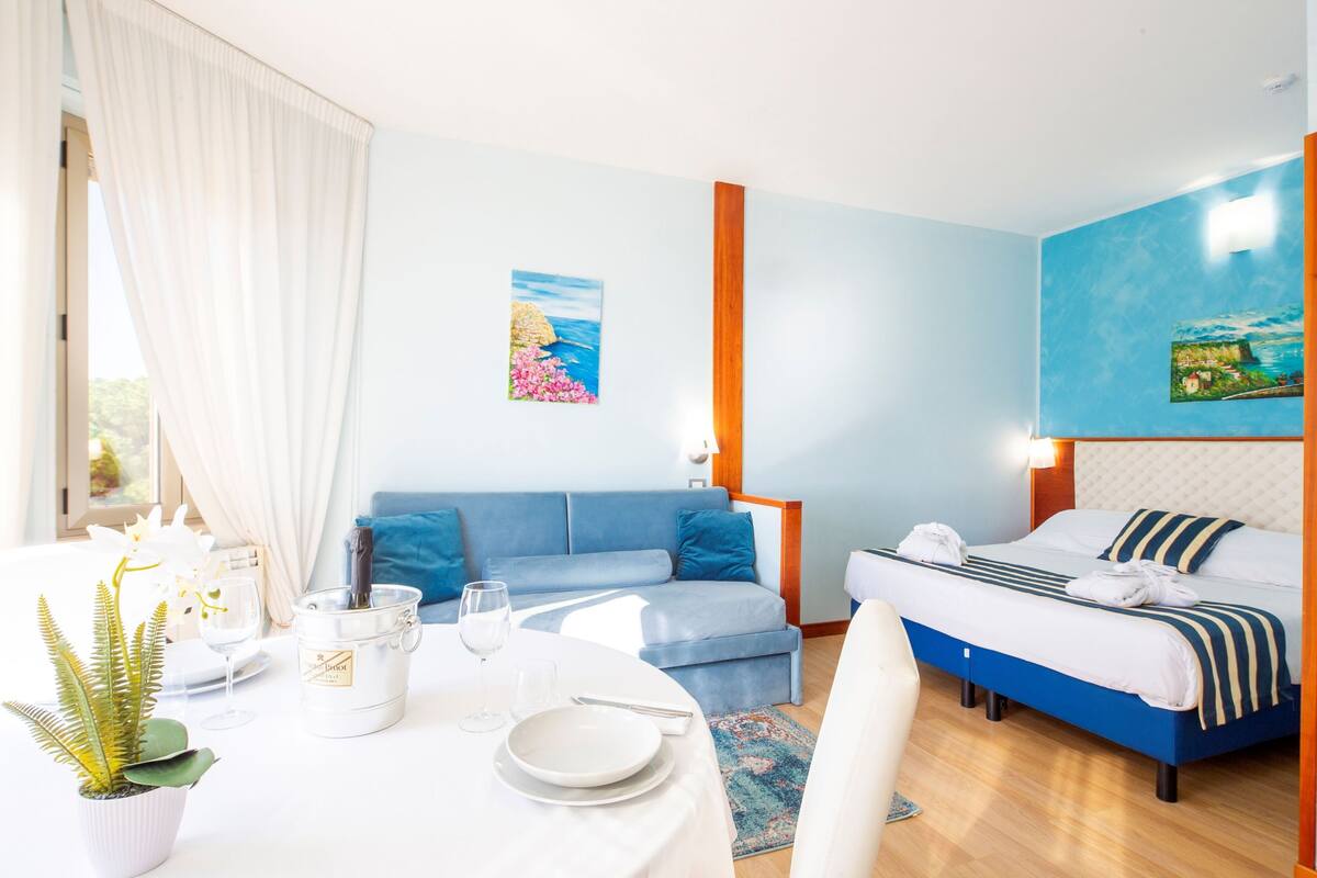 A hotel room with a bed, a sofa, a dining table with plates and a wine glass, and a potted plant.