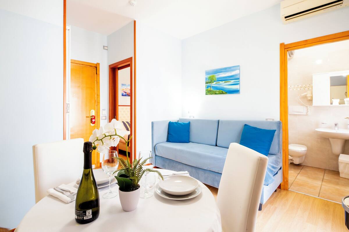 A dining area with a table set for two, a bottle of wine, a potted plant, and a blue couch in the background.