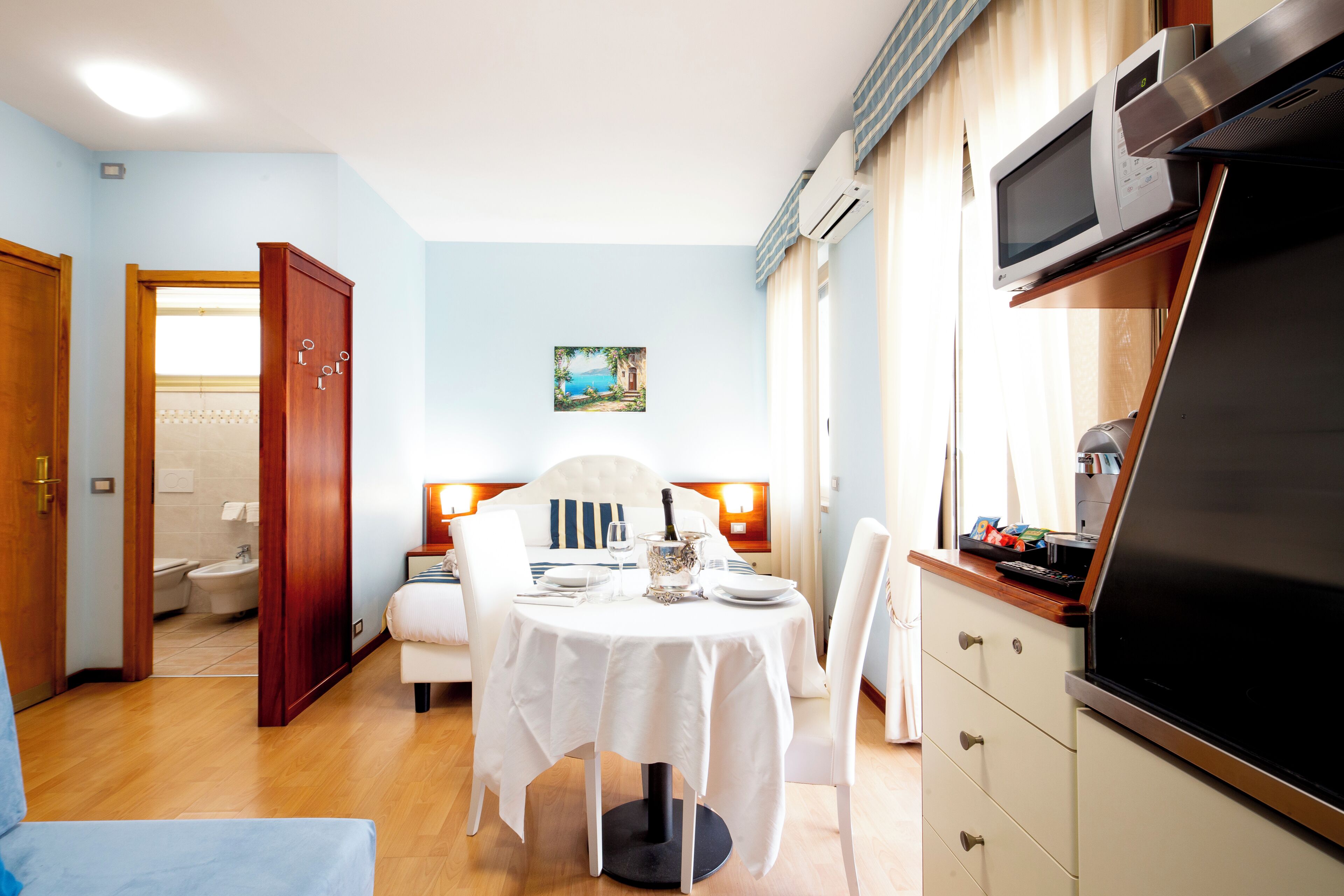 A hotel room with a dining table, white chairs, a TV, and a painting on the wall.