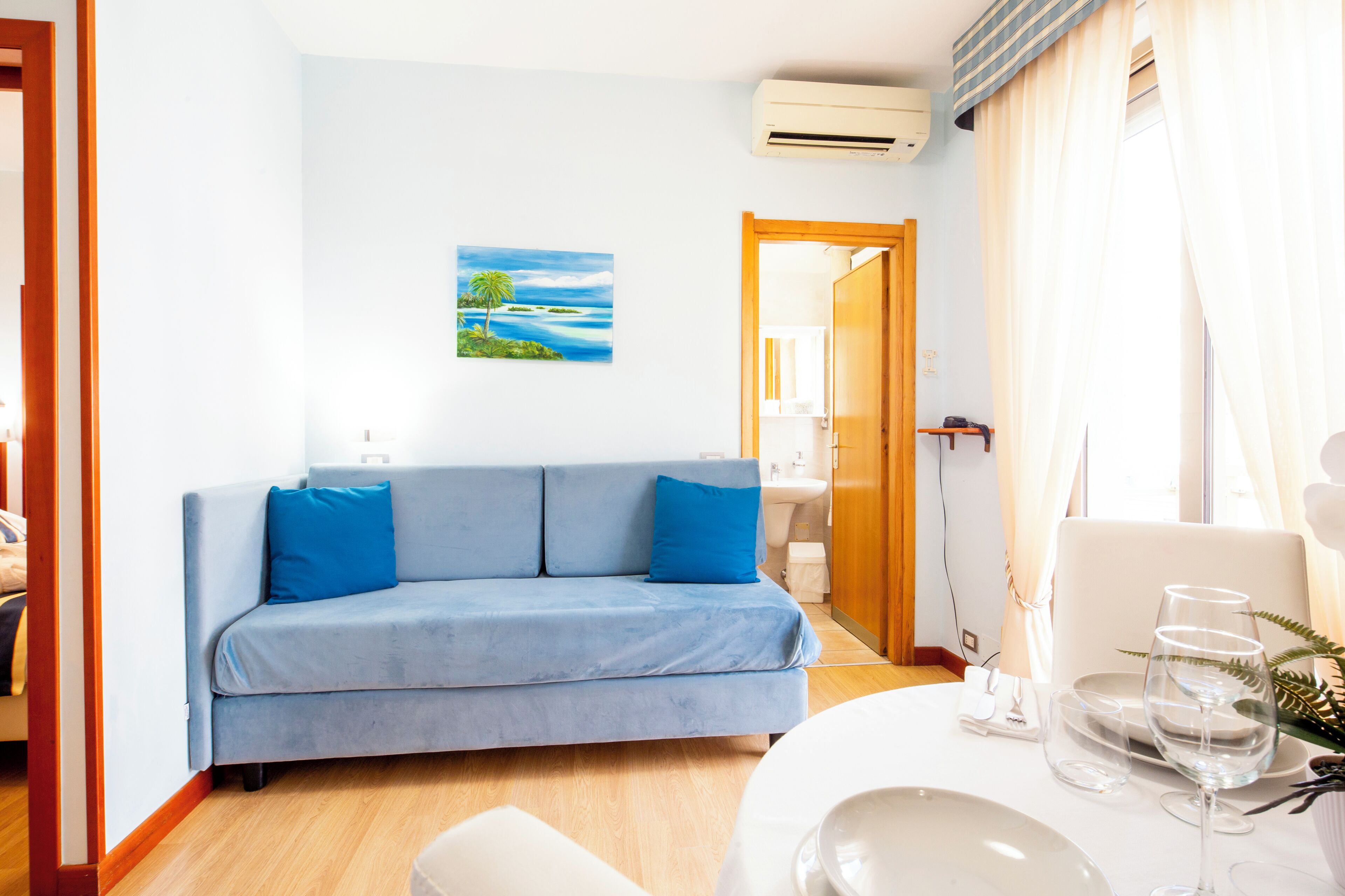 Una habitación con un sofá azul, una mesa de comedor con vasos y platos, un cuadro de un paisaje y un equipo de aire acondicionado.