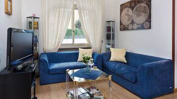 Een woonkamer met een blauwe bank, een glazen salontafel, een televisietoestel en een boekenkast.