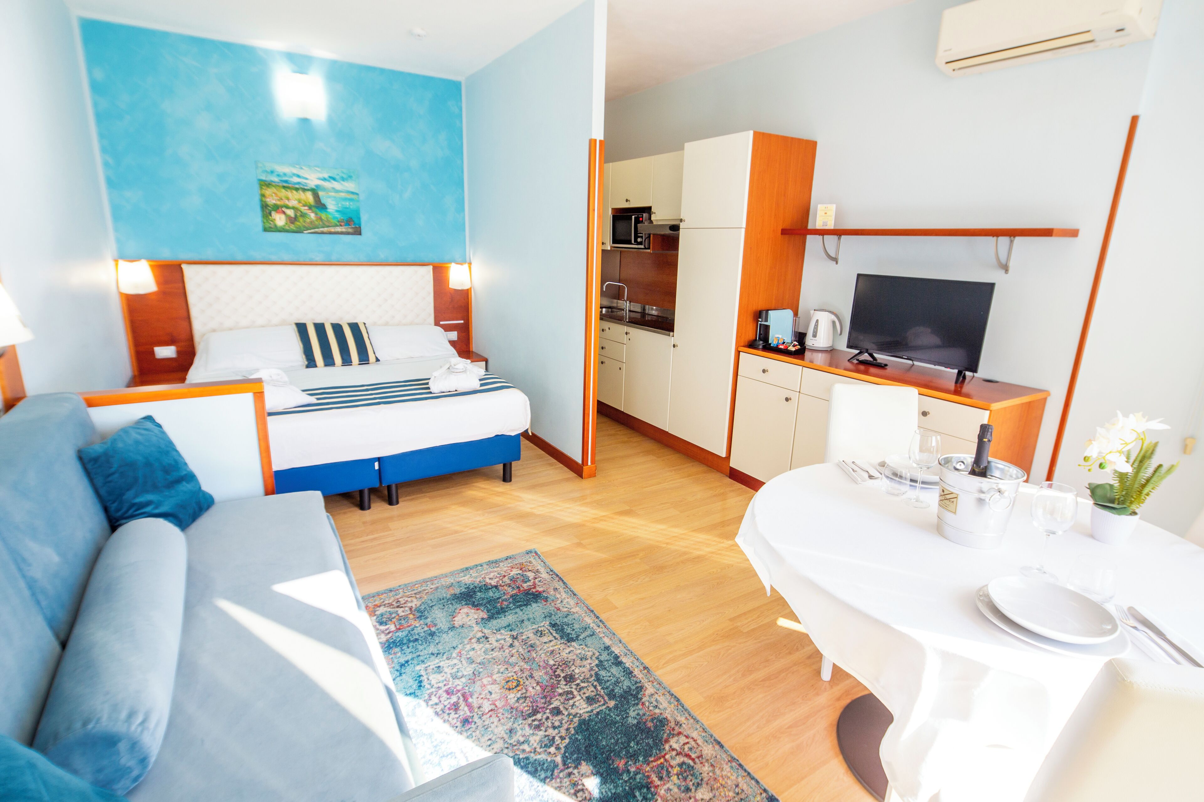 Habitación de hotel moderna con cama, sofá, mesa de comedor para dos personas, zona de cocina y televisión.