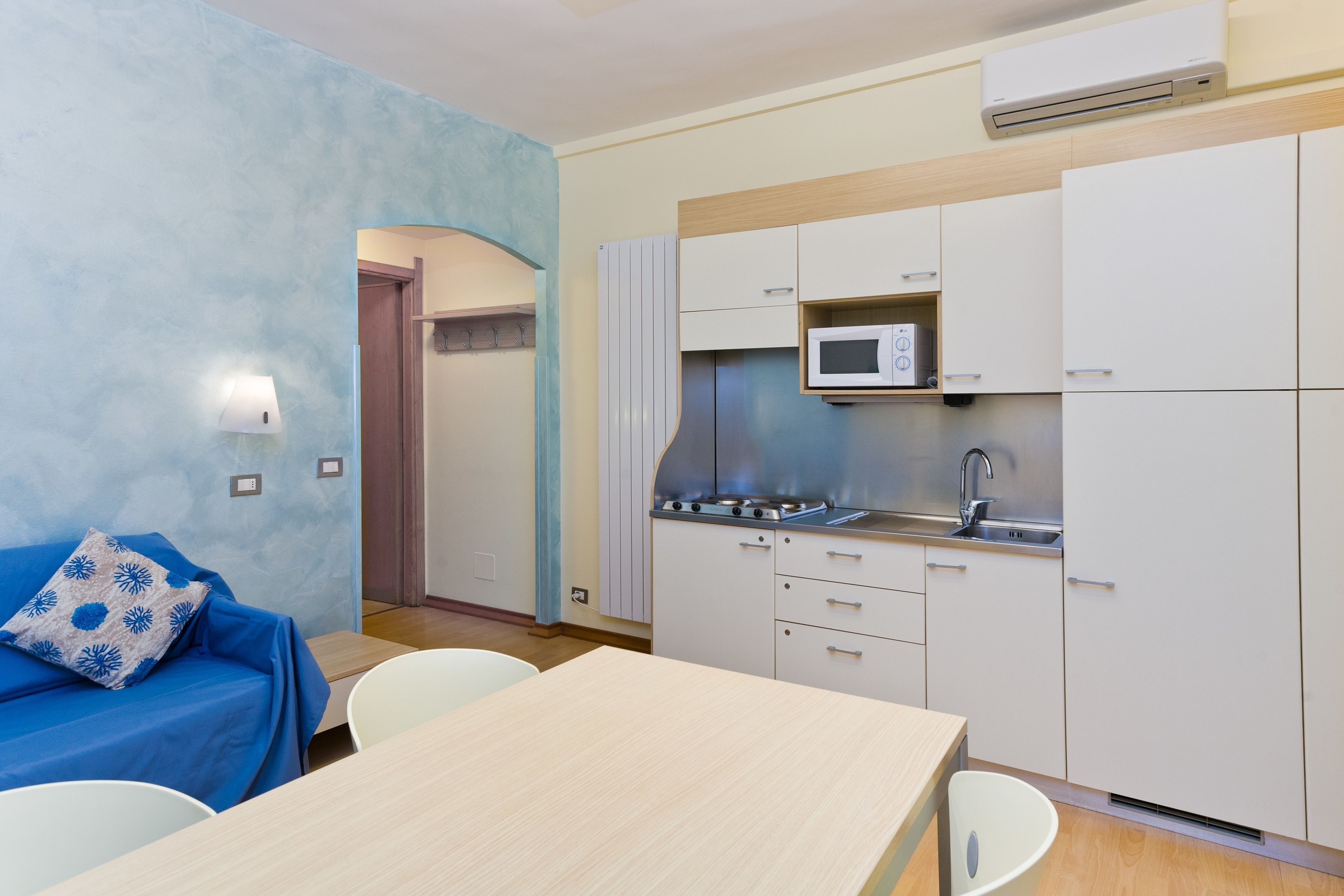 A compact kitchen with white cabinets, a microwave, and a stove. There is a dining table with chairs and a blue sofa with a patterned pillow.