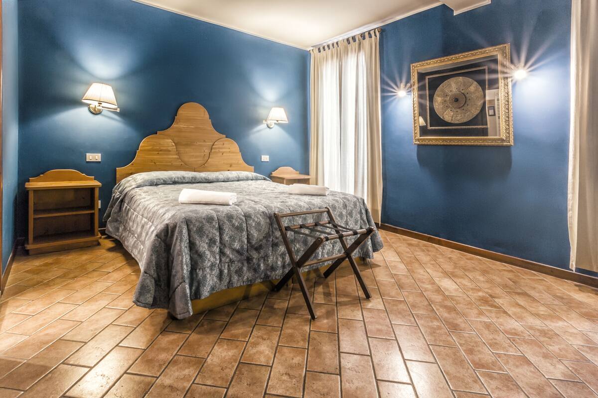 A bedroom with a wooden headboard, a bed with a gray patterned bedspread, two bedside tables with lamps, a wooden nightstand, a chair, and a framed picture on the wall.