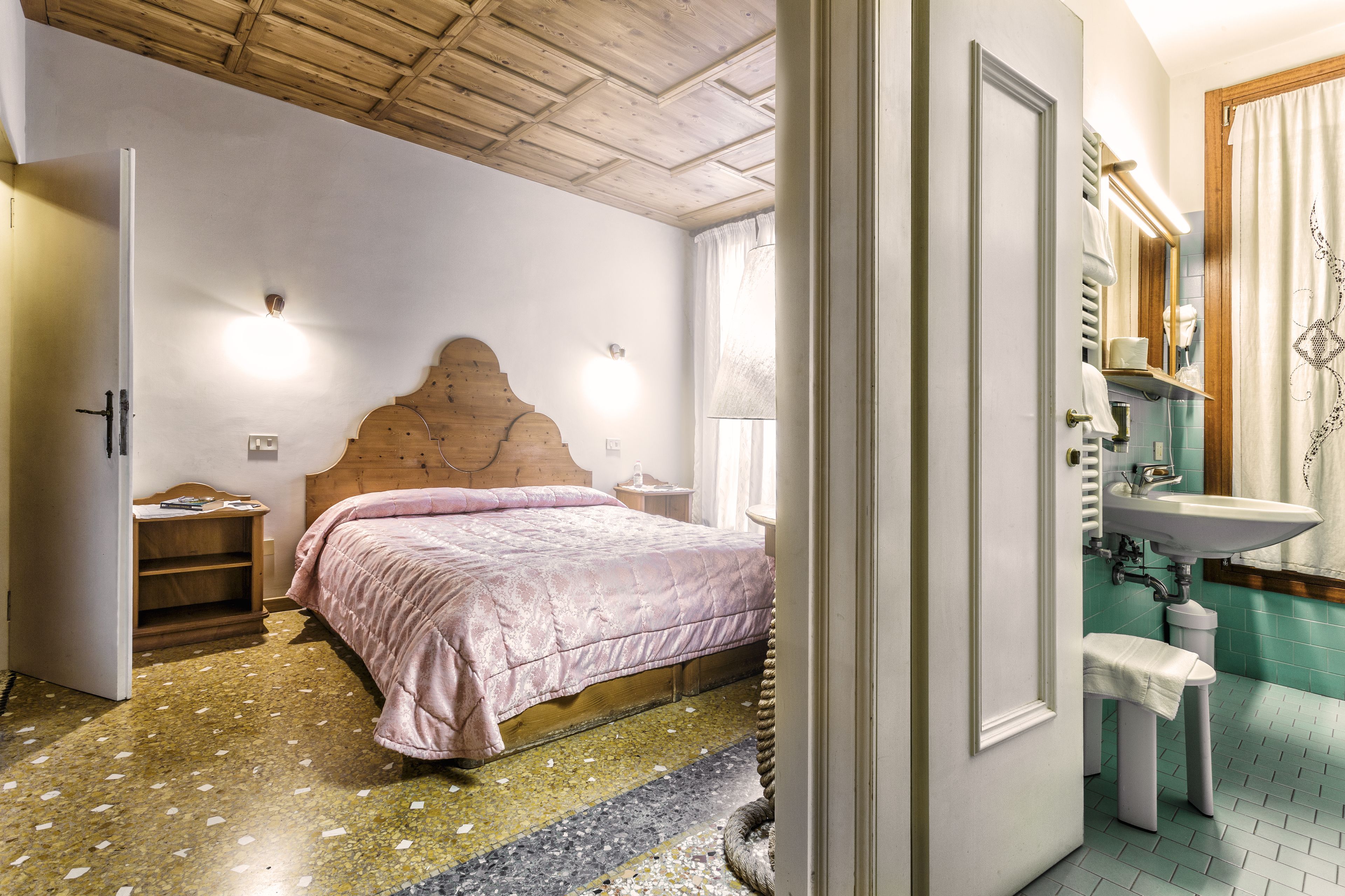 A bedroom with a wooden headboard, a bed with a pink bedspread, a nightstand, and a bathroom with a sink and mirror.
