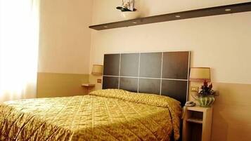 A hotel room with a large bed, a nightstand, a wall-mounted shelf, and a bedside table with a lamp.