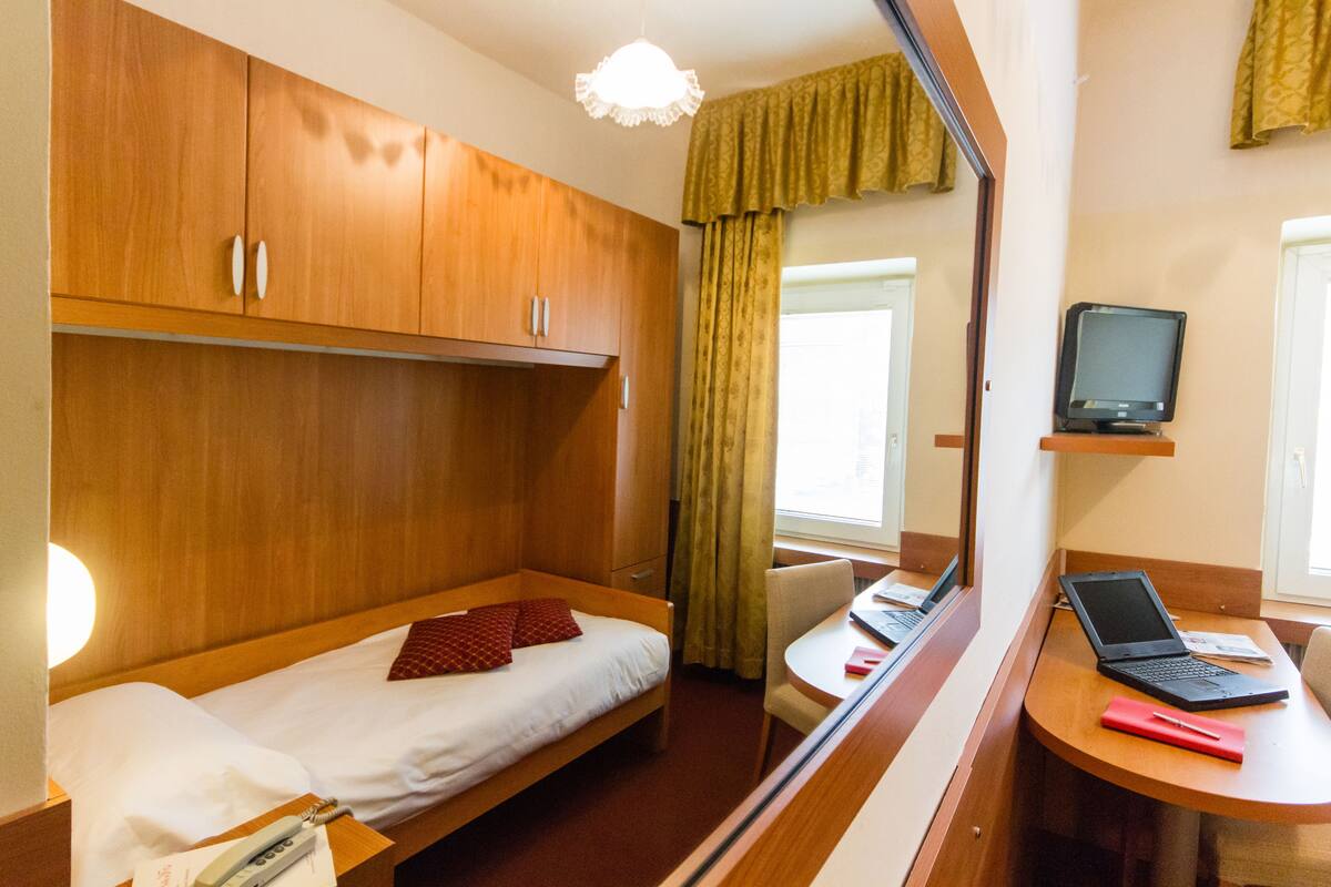 Ein kleines Hotelzimmer mit Bett, Schreibtisch, Laptop, Fernseher und Spiegel.