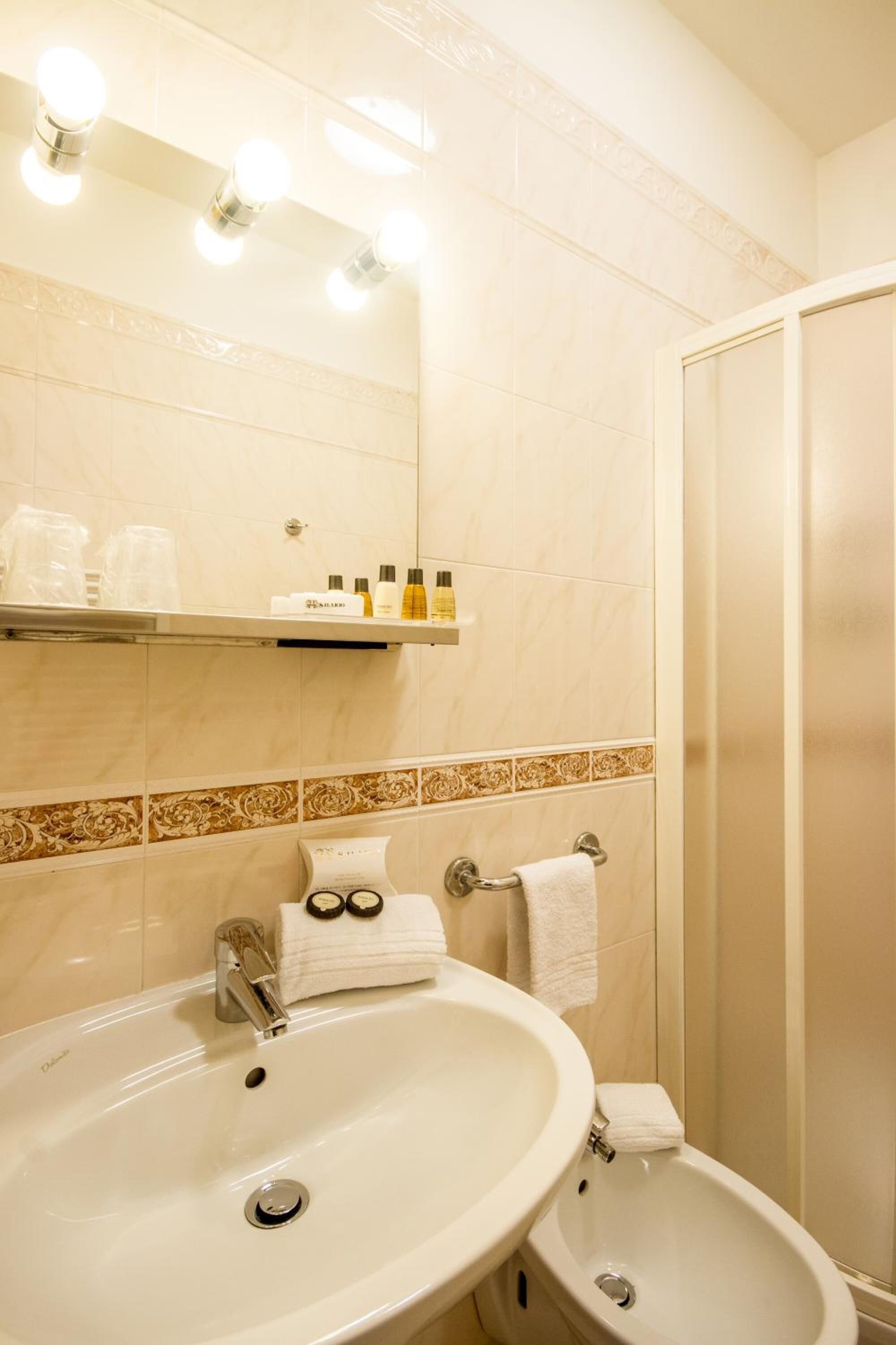 A bathroom with a large sink, a mirror, and a shower. There are toiletries on a shelf above the sink.