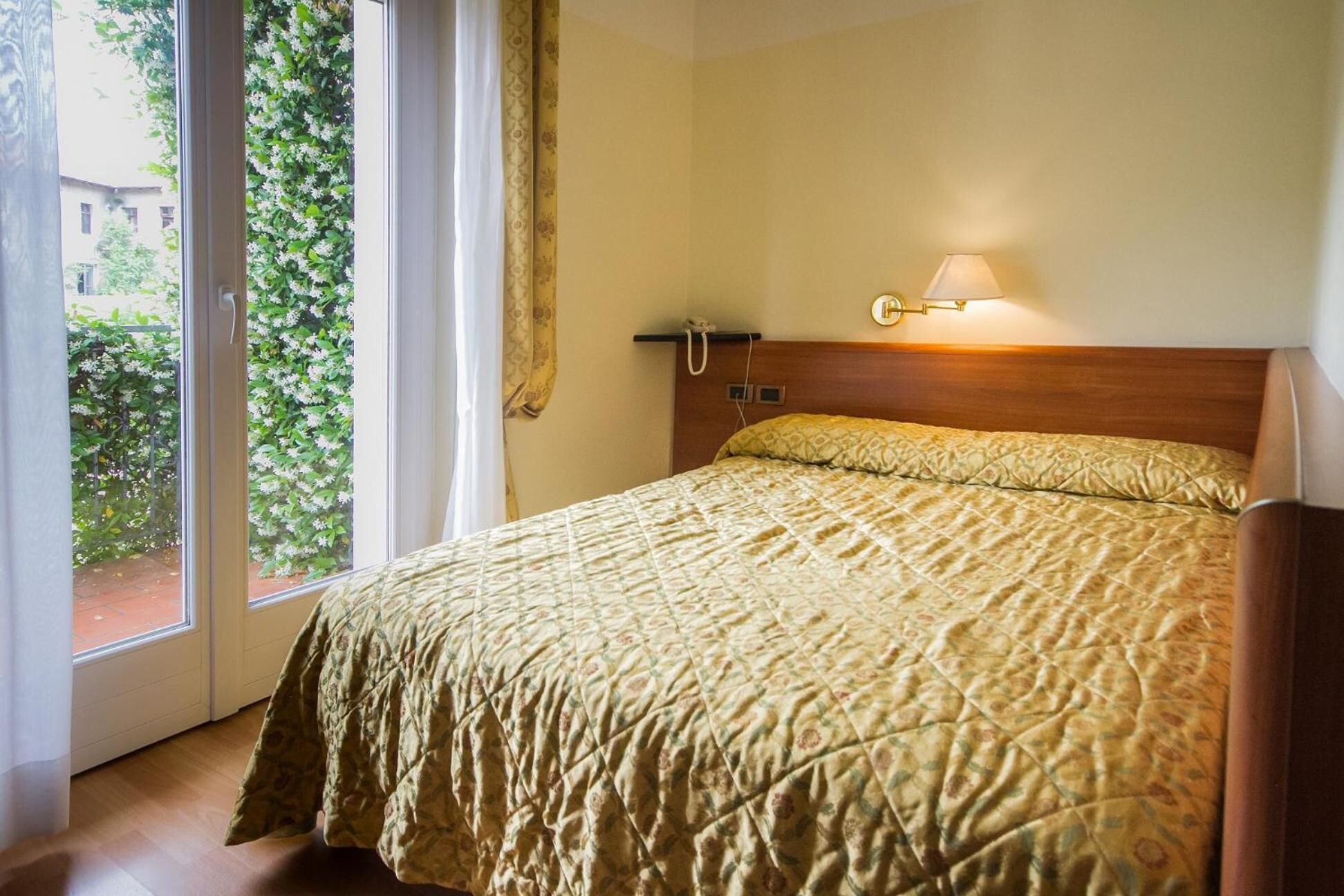 Hotellihuone, jossa on sänky, yöpöytälamppu, puhelin ja näkymä vihreälle alueelle.
