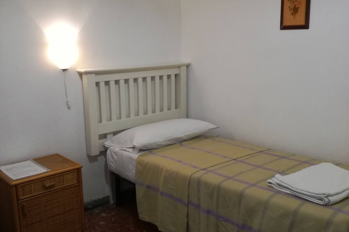 A small, simple room with a single bed, a nightstand, and a wall-mounted light.