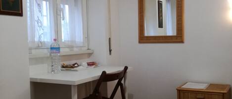 A room with a white desk, a wooden chair, a small wooden cabinet, a window with curtains, and a mirror on the wall.