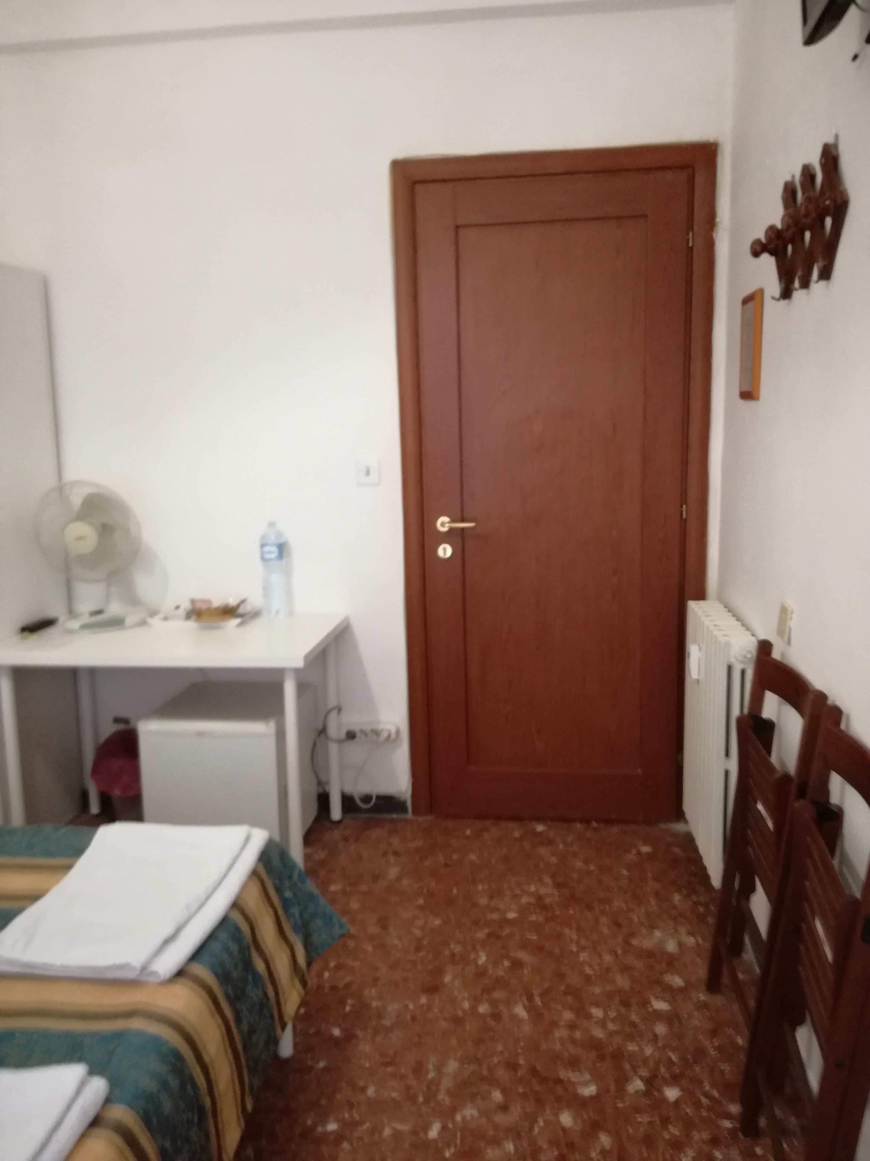 A small, simple room with a bed, a desk, a chair, a radiator, and a door.