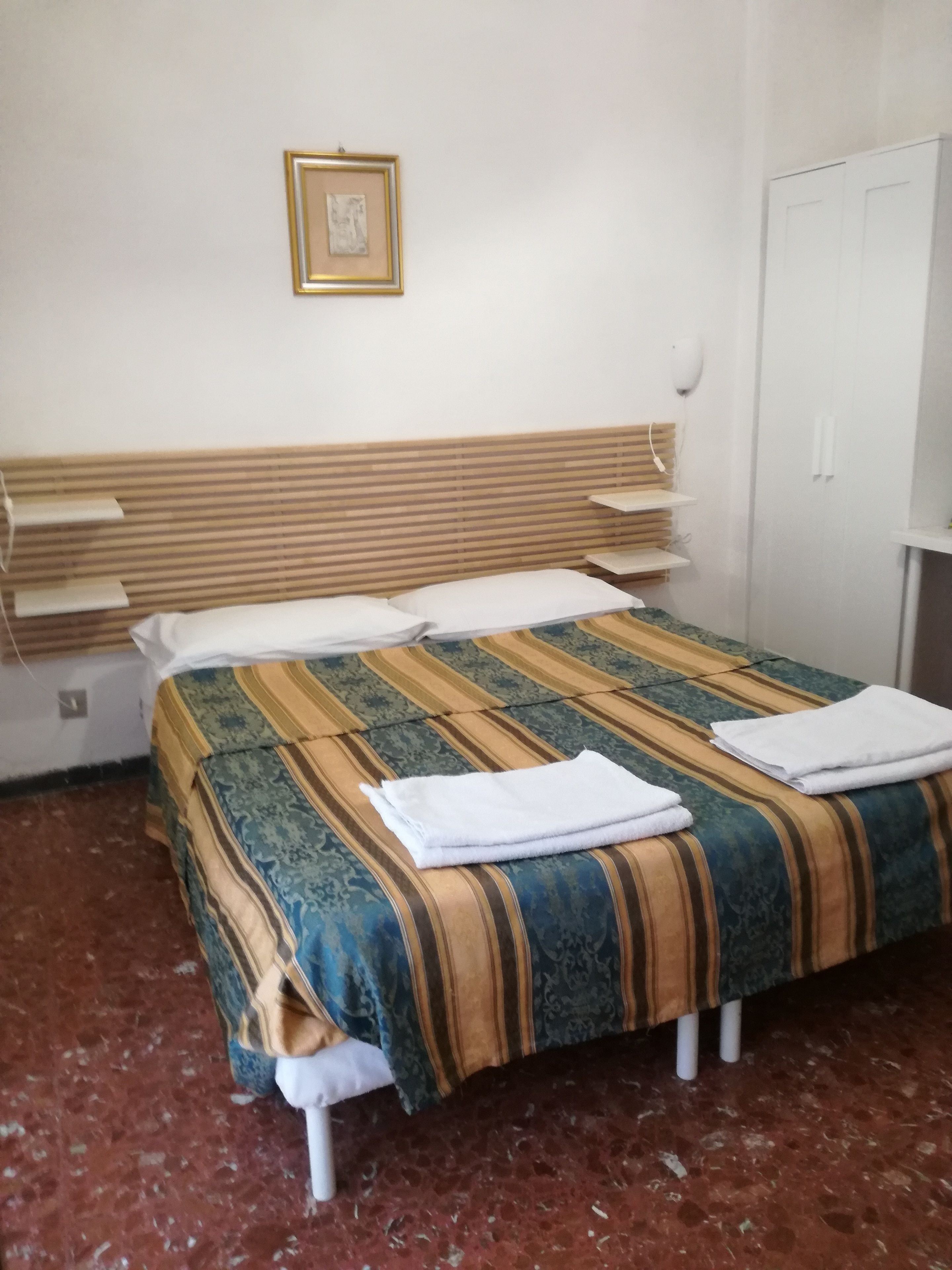 A single bed with a striped bedspread, white pillows, and a folded white towel on the bed. There is a white wardrobe to the right and a framed picture on the wall above the bed.