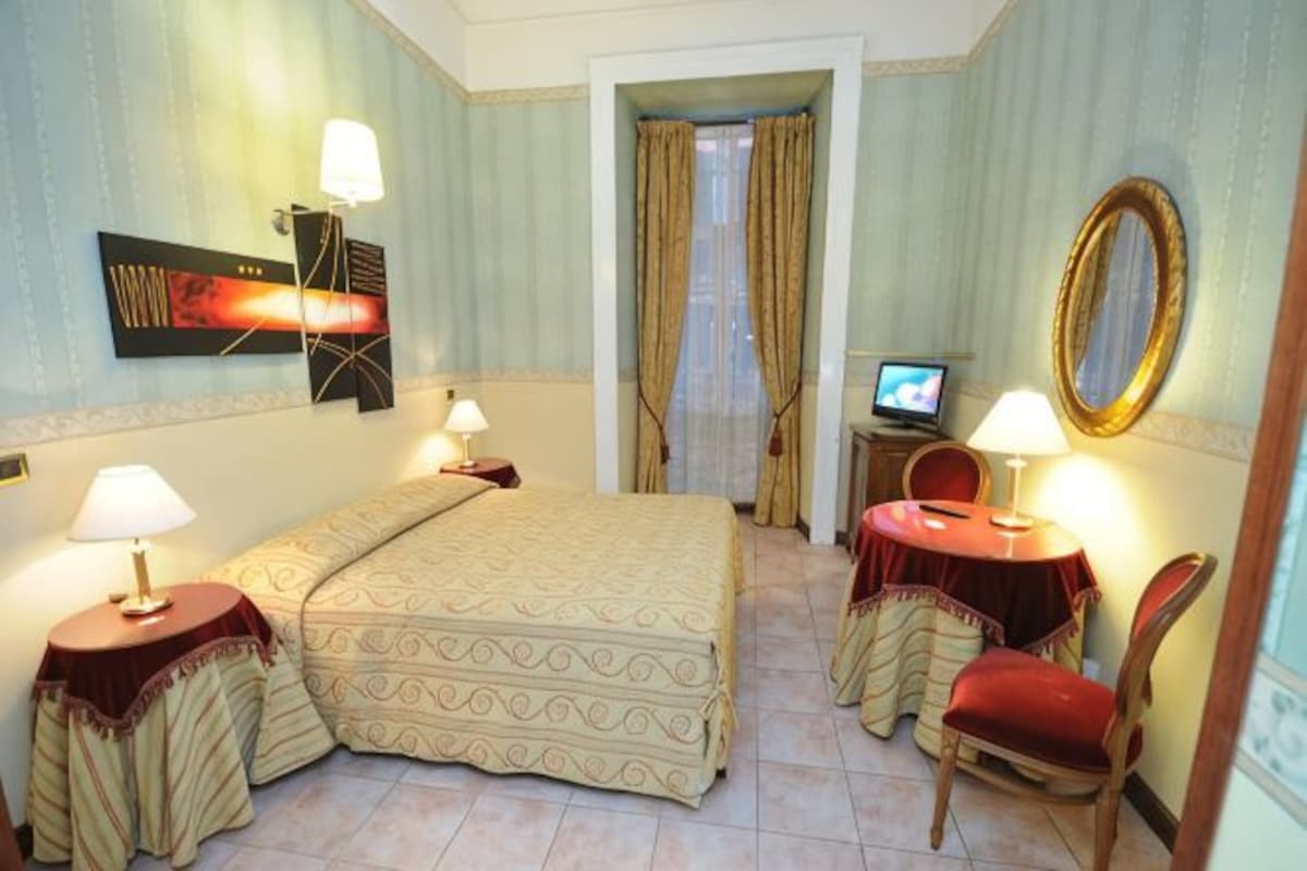 Una habitación de hotel con cama, dos mesitas de noche con lámparas, televisión, una silla y un espejo.