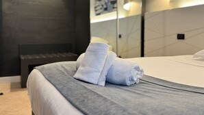 Una cama bien tendida con toallas dobladas, una pared oscura y un espejo grande que refleja la habitación.