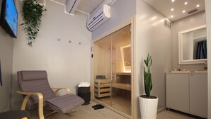 Una habitación moderna con una butaca reclinable, una planta grande en maceta, un cerramiento de vidrio corredero que da a un área de ducha y un mueble pequeño.