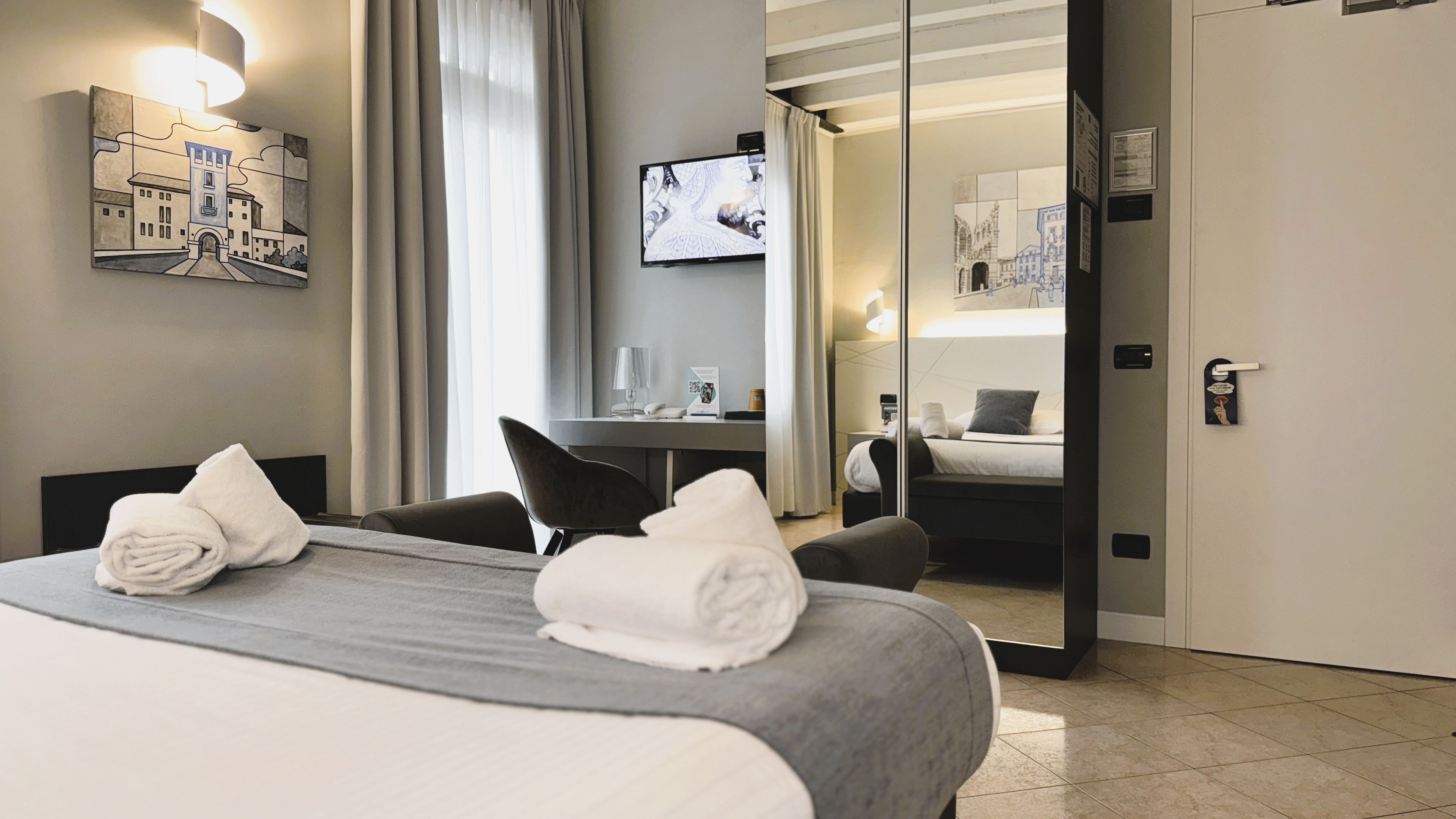 A hotel room with a bed, two towels, a desk with a lamp, a television, and a mirror reflecting the room.