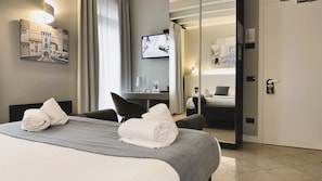 Habitación de hotel con cama, dos toallas, escritorio con lámpara, televisión y espejo que refleja la habitación.