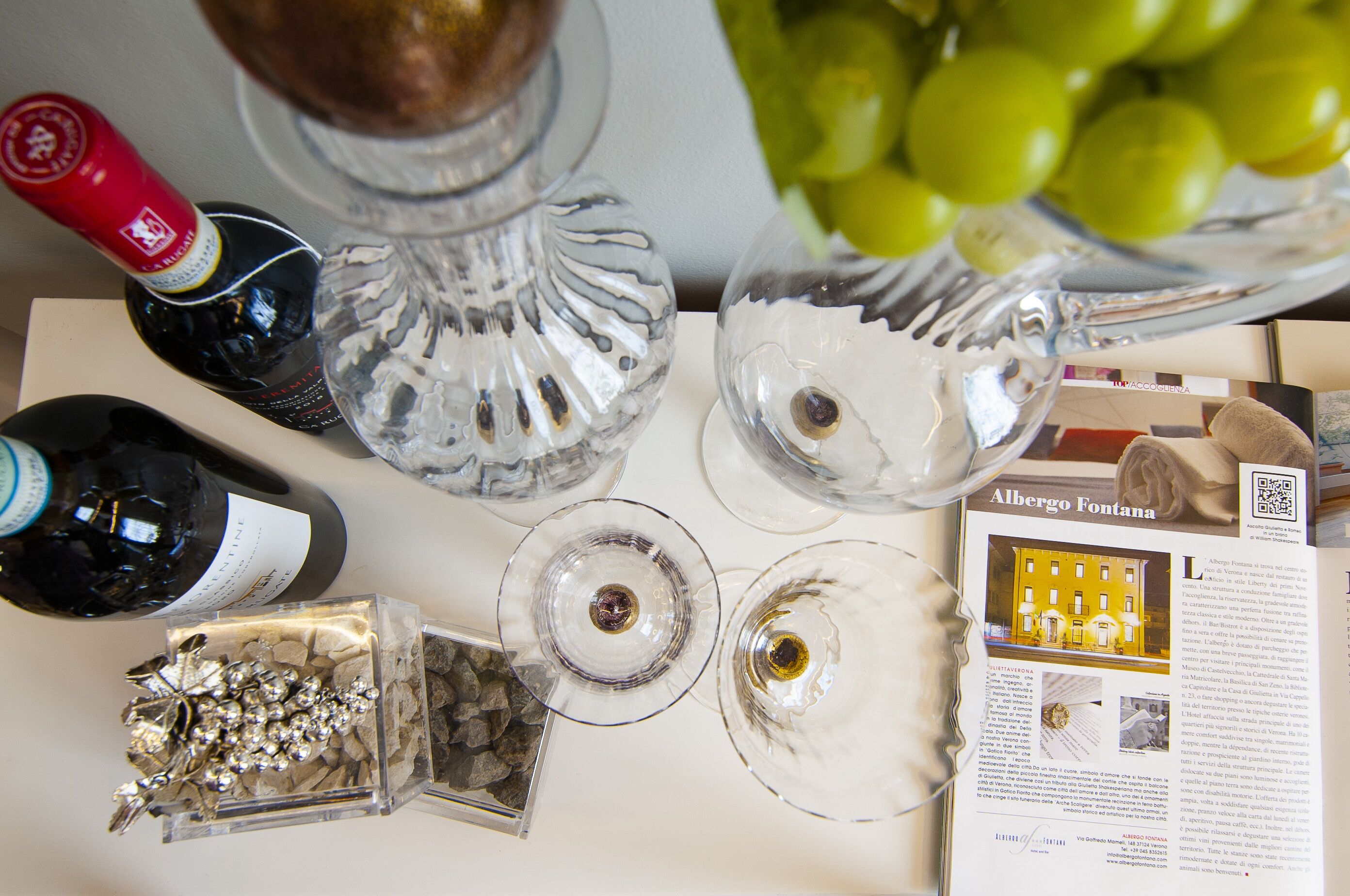 A table with wine bottles, glasses, a decorative silver branch, a bowl of grapes, and a magazine featuring a building.