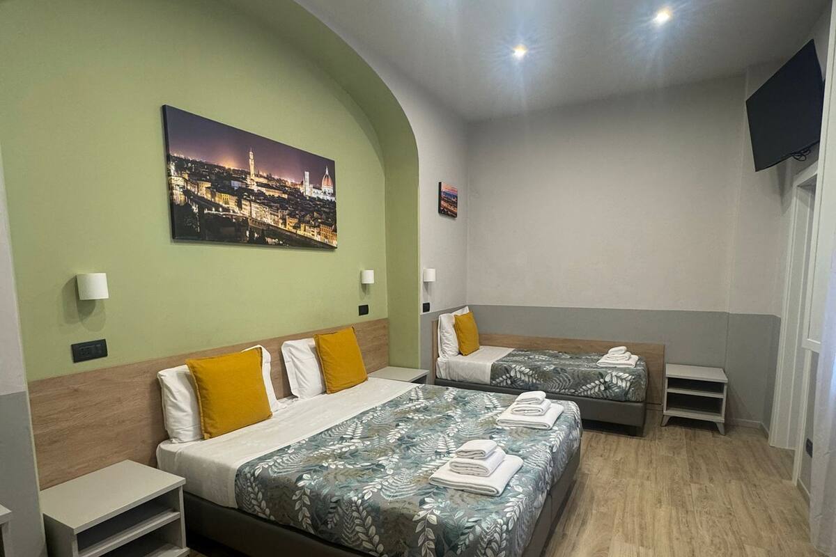 Una habitación de hotel con una cama grande, una cama más pequeña, un televisor y un cuadro con una vista urbana en la pared.