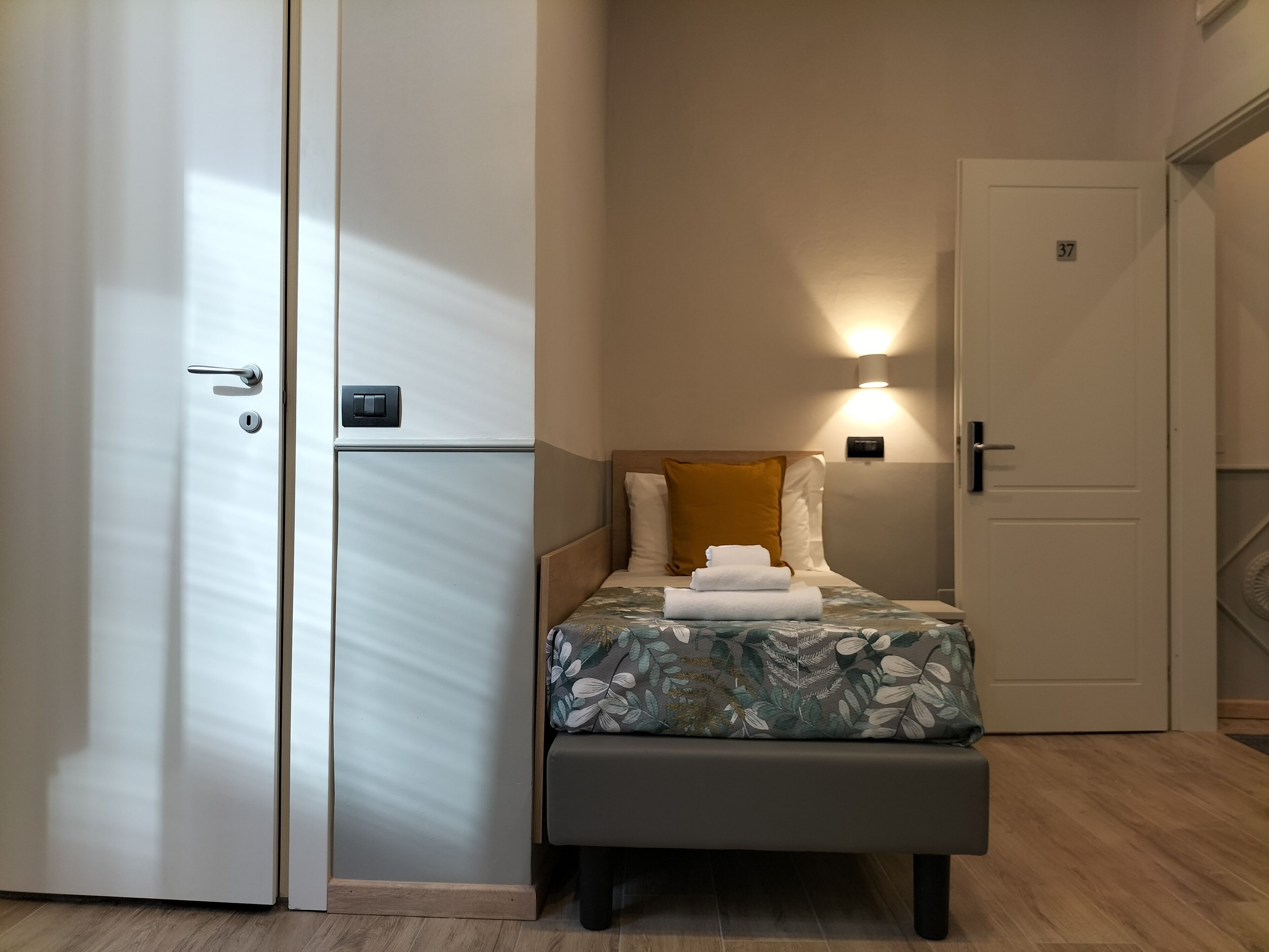 Habitación de hotel pequeña con cama, mesita de noche, lámpara de pared y puerta con número.