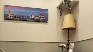 A hotel lobby with two white armchairs, a lamp, a framed cityscape painting, and a small table with a vase of flowers. - Hotel Camilla Guelfa (Florence)