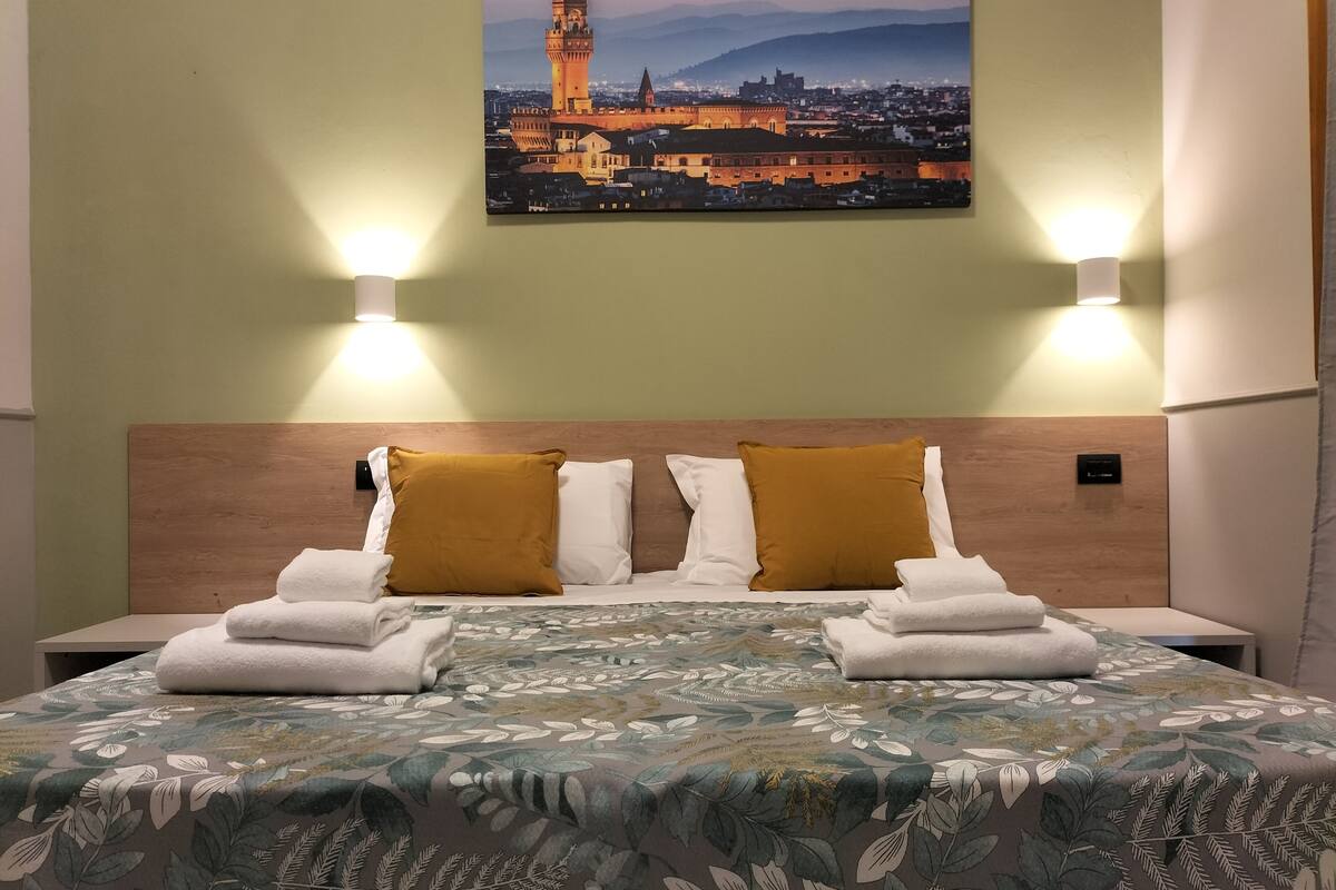 Habitación de hotel con cama, dos almohadas, una mesita de noche y un cuadro de paisaje urbano colgado en la pared.