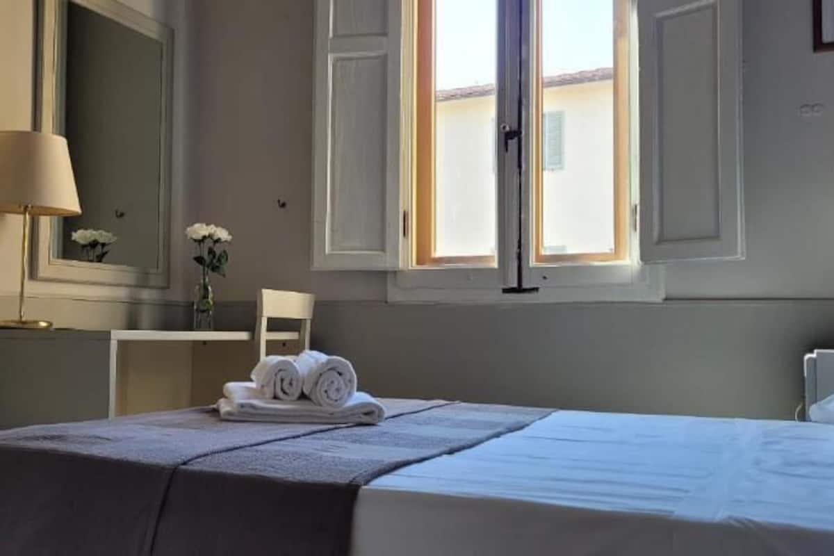 Una habitación de hotel con cama, escritorio, silla, lámpara y ventana con cortinas.