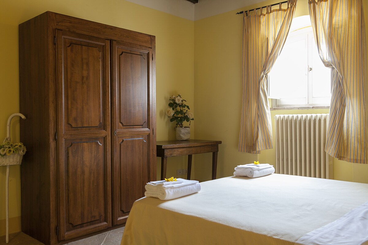 Quarto de hotel com cama, guarda-roupa de madeira, uma mesinha com um vaso de flores e uma janela com cortinas.