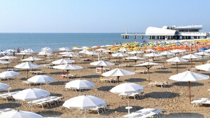 Una playa con hileras de sombrillas blancas y tumbonas, un muelle con un edificio blanco, y el mar al fondo.