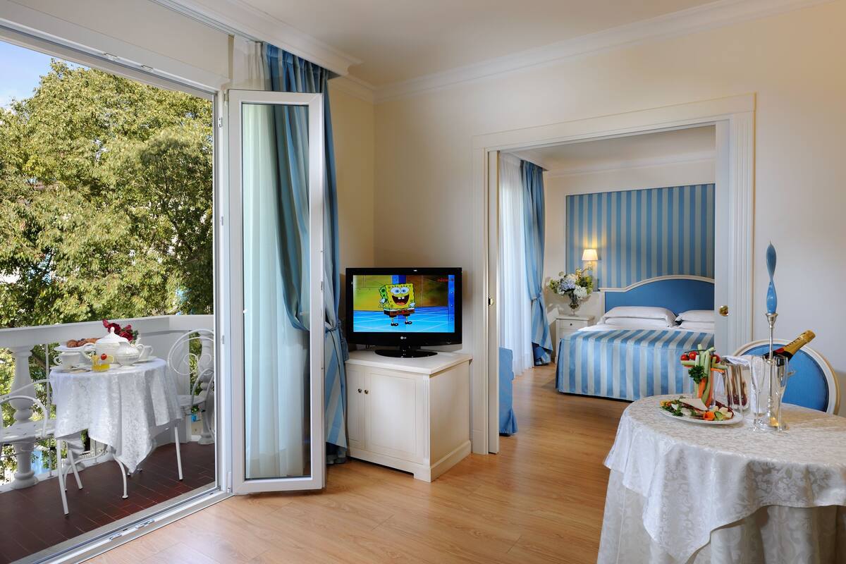 Ett hotellrum med en balkong, en tv, en säng med blå- och vitrandigt sänglinne, ett matbord med en sallad och ett champagneglas.